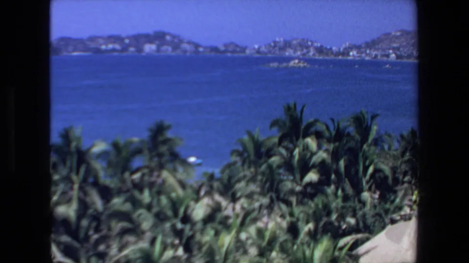 1972 coastal home-movie frame from Mazatlan showing palm tops, open blue harbor water, and a built shoreline at the base of hills.