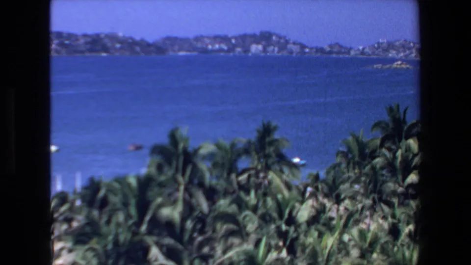 1972 Mazatlan vacation footage looking across a blue coastal bay toward distant hills, with palm trees and small boats visible near the shore.