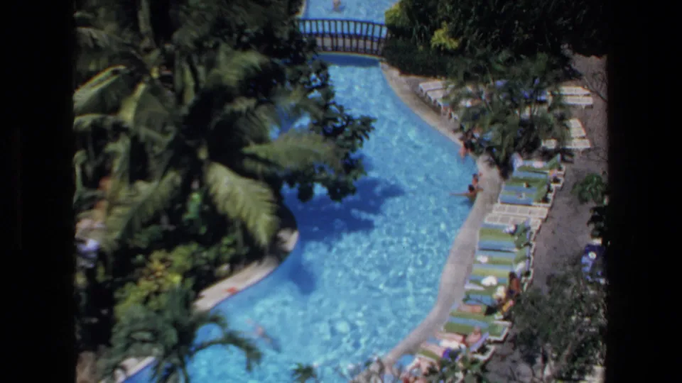 1972 Mazatlan resort scene showing a winding outdoor swimming pool bordered by lounge chairs, palms, dense greenery, and a few visible guests.
