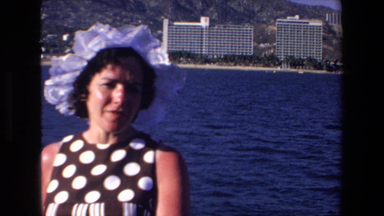 In Mazatlan, Mexico in 1972, a woman in a large white bonnet and dark polka-dot dress faces the camera near the ocean, with beachfront resort hotels and hills in the background.