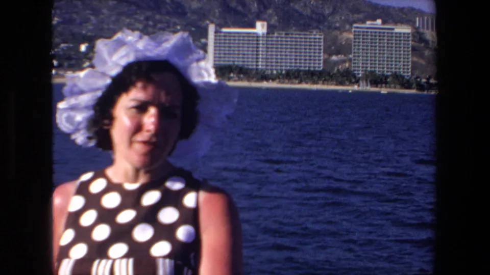 In Mazatlan, Mexico in 1972, a woman in a large white bonnet and dark polka-dot dress faces the camera near the ocean, with beachfront resort hotels and hills in the background.
