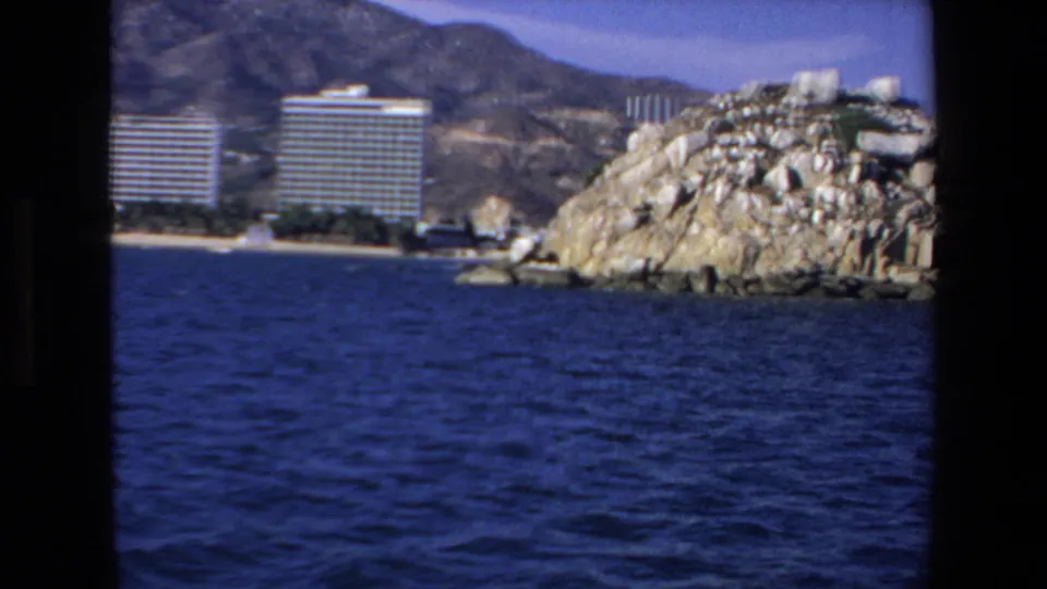 The camera frames Mazatlan shoreline hotels beside a rocky coastal point, with deep blue water filling the foreground and hills beyond.