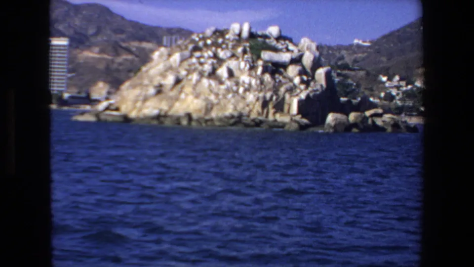 A rocky Mazatlan coastal formation rises from the shoreline, with blue bay water in the foreground and resort structures partly visible at the edge of the frame.