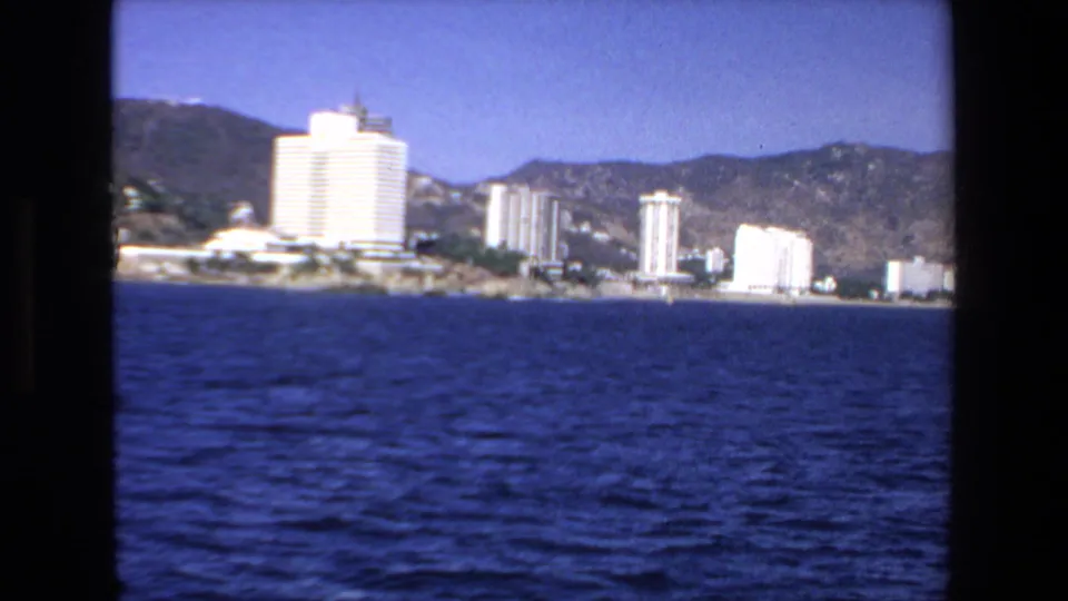 Mazatlan, Mexico in 1972, with beachfront towers and mountain slopes viewed from across deep blue water in 8mm footage.