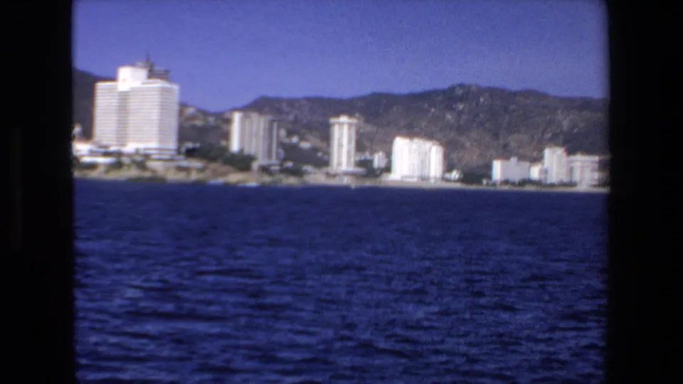A 1972 8mm view of Mazatlan's developed shoreline, showing pale high-rise buildings below brown-green mountains across open water.