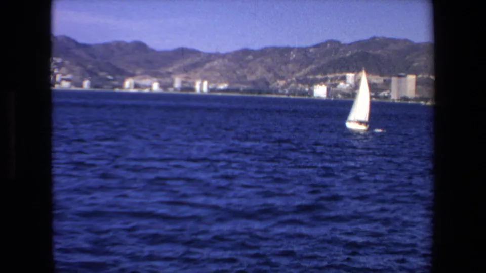A small white sailboat crosses the foreground off Mazatlan in 1972, with the city shoreline and mountains visible in the distance.