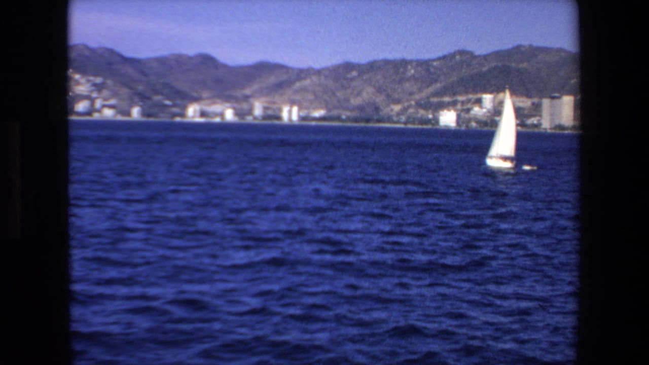 1972 Mazatlan home-movie frame showing a white sailboat crossing blue water with shoreline buildings and rugged mountains in the background.