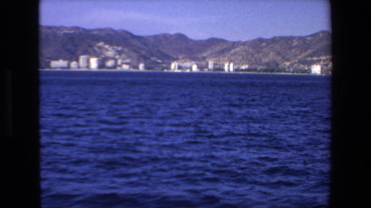 Blue ocean in foreground with distant shoreline buildings and mountains under clear sky.