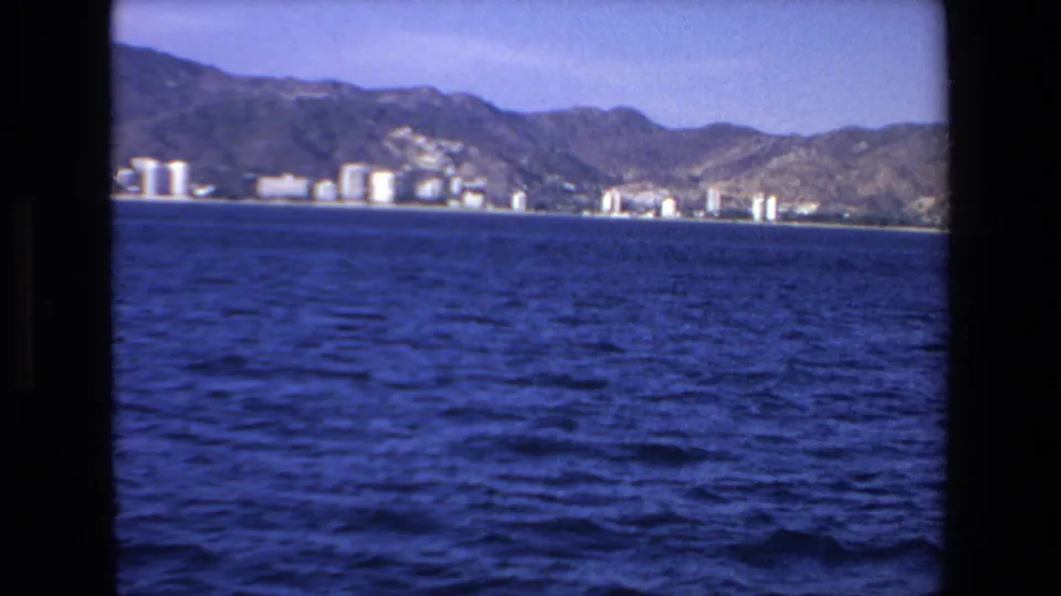 Soft 8mm frame of the Mazatlan shoreline seen across open water, with buildings clustered below a mountain ridge.
