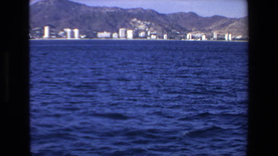 Wide 1972 Mazatlan coastal frame showing choppy blue water in the foreground, a line of waterfront buildings, and mountains behind them.