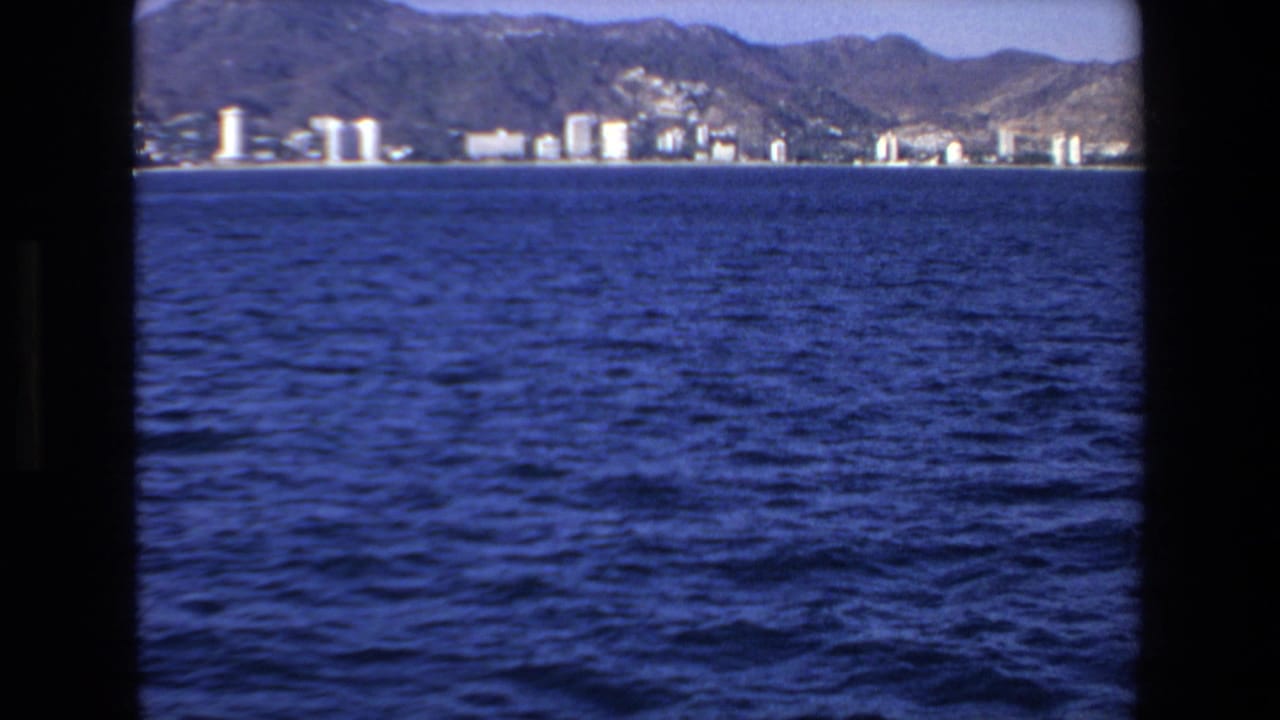 1972 8mm frame of Mazatlan, Mexico, with blue water in the foreground, a distant beachfront skyline, and mountains behind the shore.