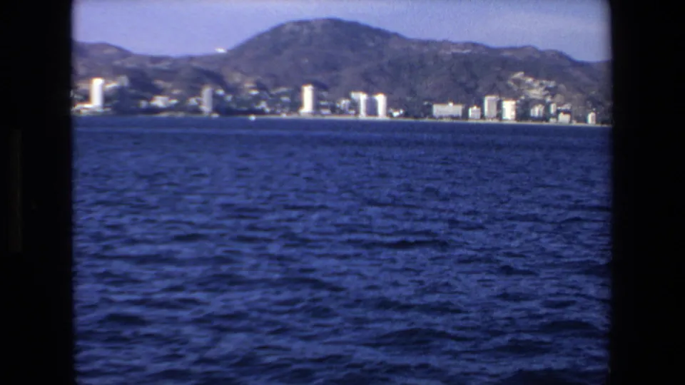 Soft 8mm view of Mazatlan from offshore, showing choppy blue water, scattered waterfront buildings, and a mountainous backdrop.