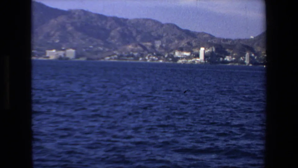 Final 1972 8mm frame looking toward Mazatlan from the boat, with open water, a distant coastal strip, tall buildings, and rugged mountains.