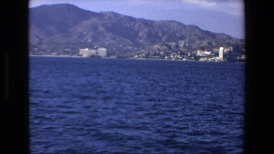 1972 Mazatlan, Mexico 8mm frame showing open blue water in the foreground and a distant town backed by rugged mountains.