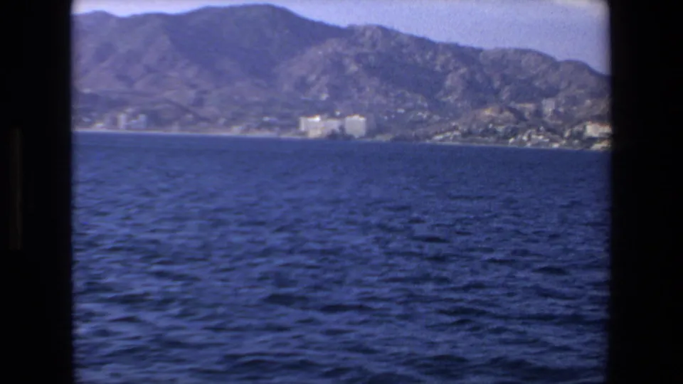 1972 Mazatlan coastal view from a moving boat, with rippled ocean water, distant buildings, and mountains behind the shoreline.