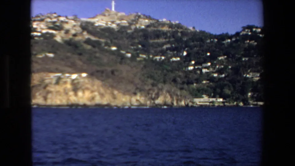 1972 Mazatlan 8mm frame showing a closer rocky shoreline with hillside buildings, trees, and a prominent white cross near the top of the ridge.