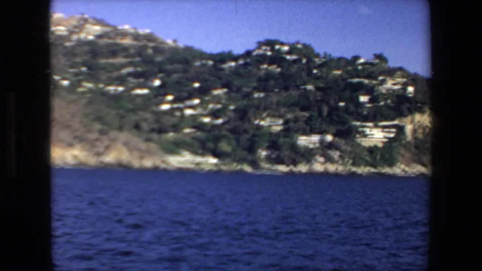 1972 Mazatlan shoreline from the boat, with blue water below a rocky coast and white hillside buildings among dark green vegetation.