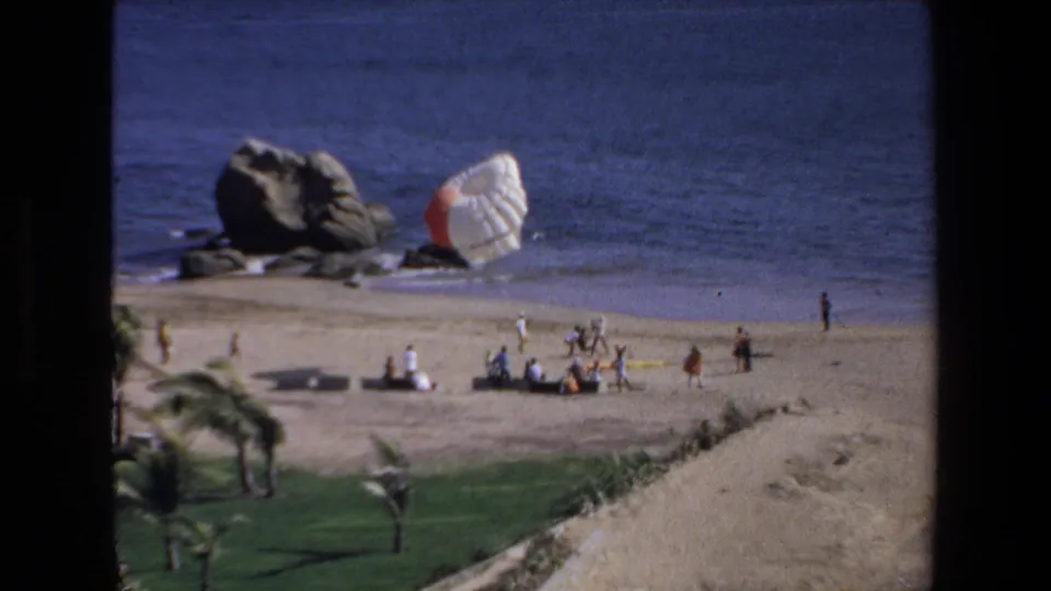 Mazatlan, Mexico 1972 8mm frame showing a sandy beach, ocean, rocky outcrop, and people watching a large white parachute near the shoreline.