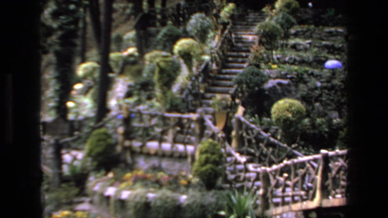 1972 Mazatlan travel footage frame showing a landscaped garden area with stone steps, trimmed shrubs, tall trees, and rustic railings.
