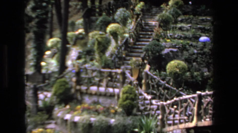 1972 Mazatlan travel footage frame showing a landscaped garden area with stone steps, trimmed shrubs, tall trees, and rustic railings.