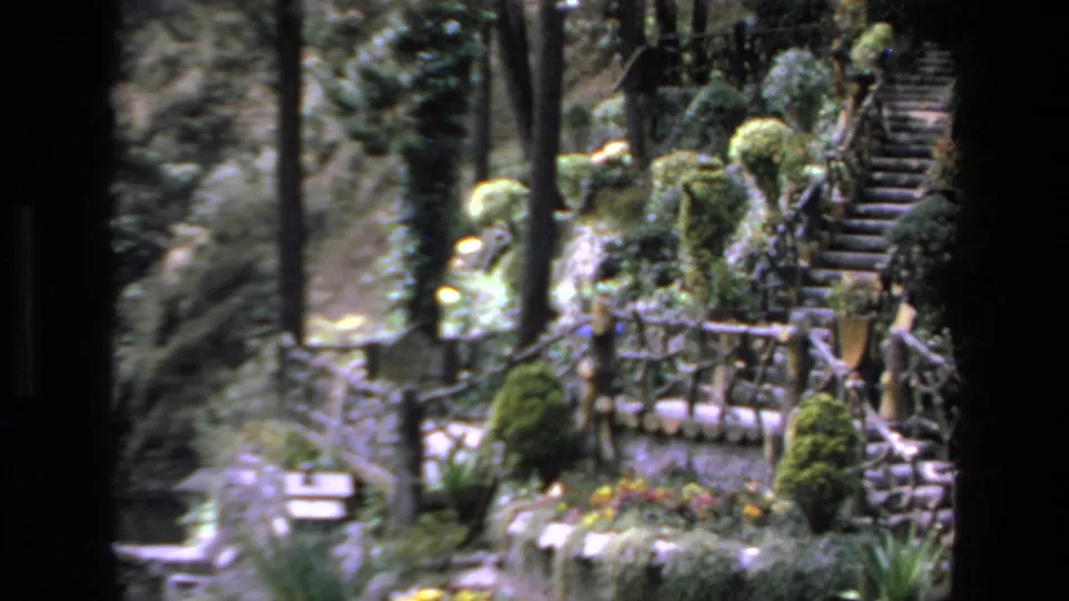 Mazatlan, Mexico 1972 8mm frame of a lush garden terrace with stonework, flowering plants, clipped shrubs, wooden railings, and steps leading upward.