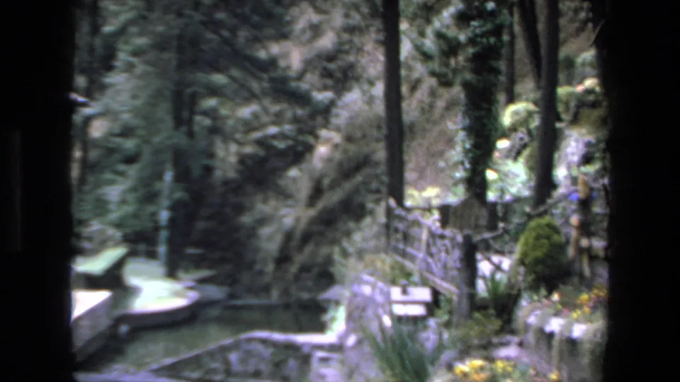 1972 Mazatlan home-movie frame showing a garden walkway with rustic railing, dense greenery, clipped shrubs, and a small pool or fountain edge.