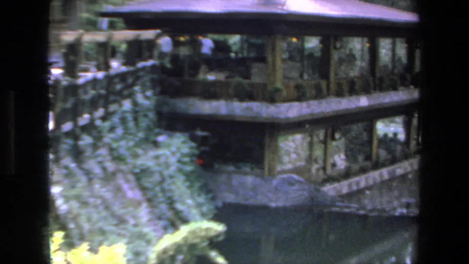 1972 Mazatlan home-movie frame showing an open-air cabana or pavilion beside water, with trimmed hedges, garden plantings, and small background figures.