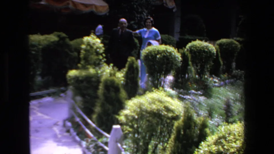 1972 Mazatlan home-movie frame showing a couple strolling together along a landscaped path among rounded hedges and dense resort garden plantings.