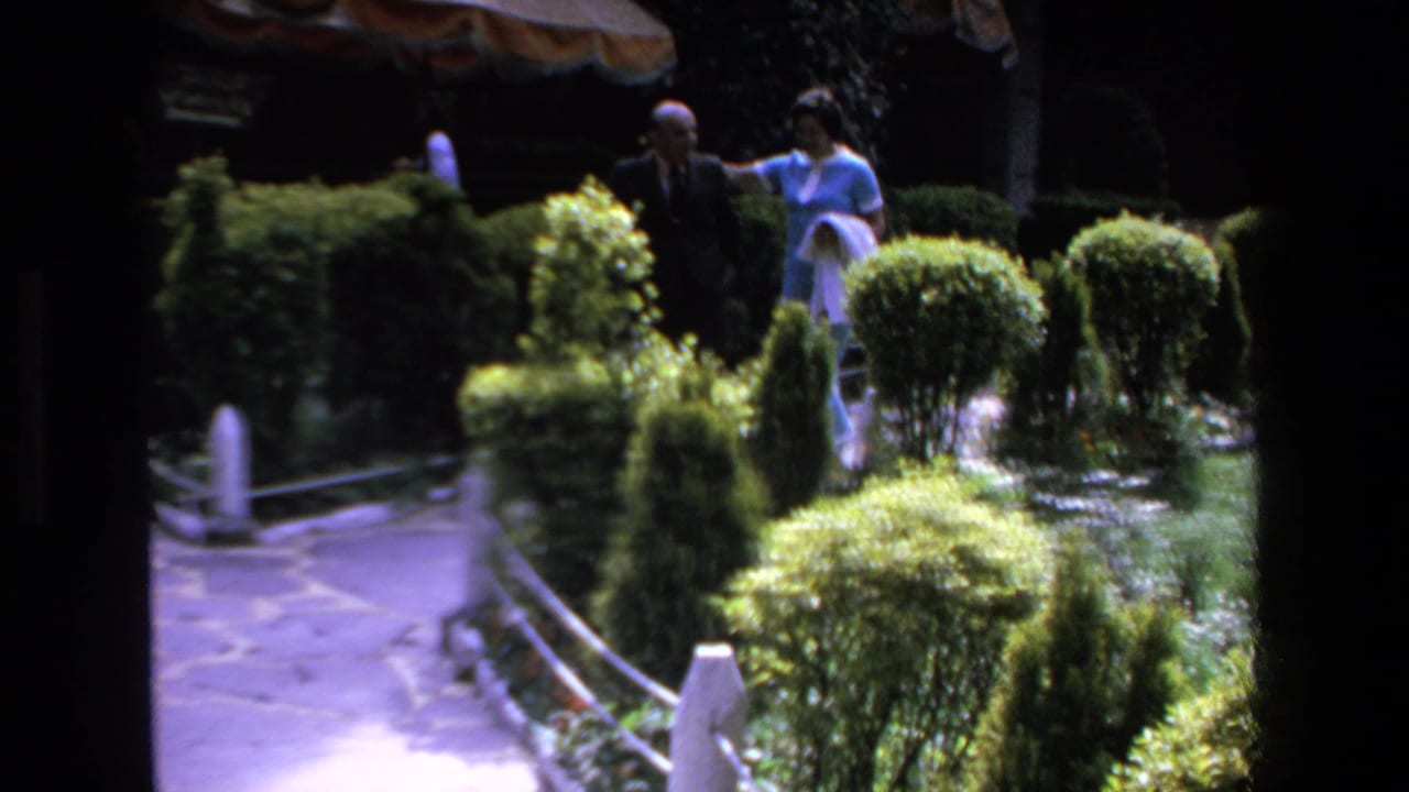 In Mazatlan, Mexico in 1972, a man in a dark suit and a woman in a light blue outfit appear near trimmed hedges along a stone garden path.