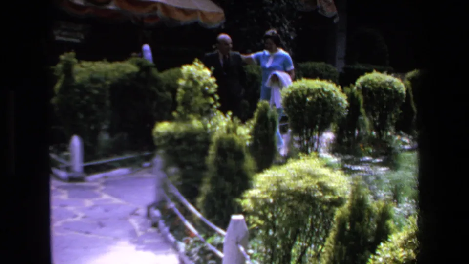 In Mazatlan, Mexico in 1972, a man in a dark suit and a woman in a light blue outfit appear near trimmed hedges along a stone garden path.