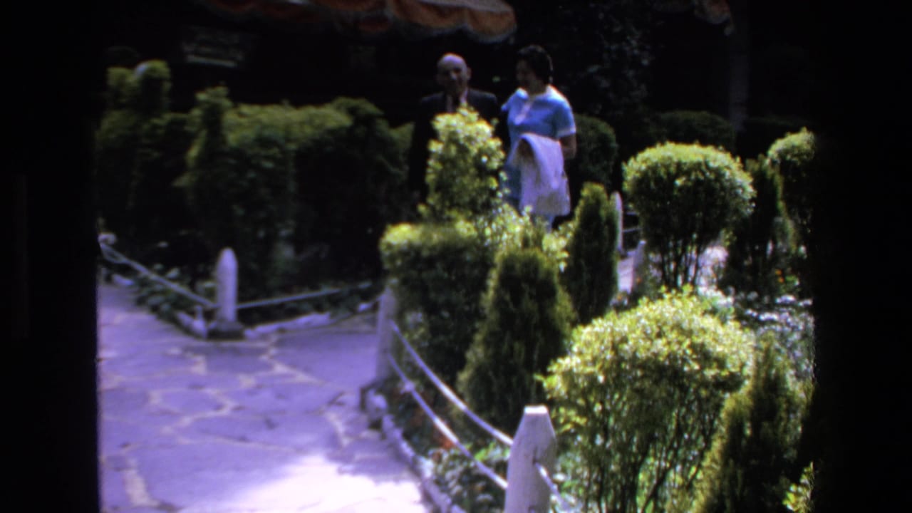 Two adults stand or walk near hedges and a shaded garden path.