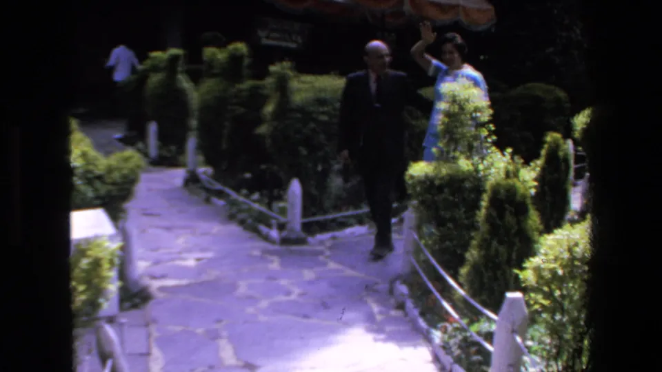 A suited man and a woman in a light blue outfit walk toward the camera on a stone path in Mazatlan, with the woman raising one arm as hedges line the walkway.