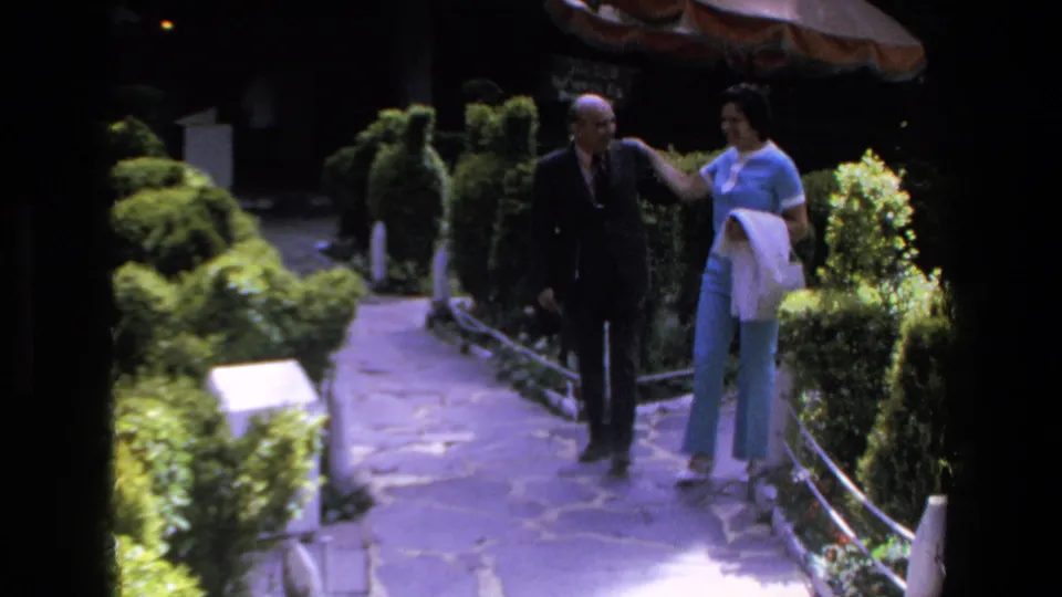 The man and woman walk arm-in-arm along the manicured garden path in the supplied 1972 Mazatlan footage, with bright foliage and white railings around them.