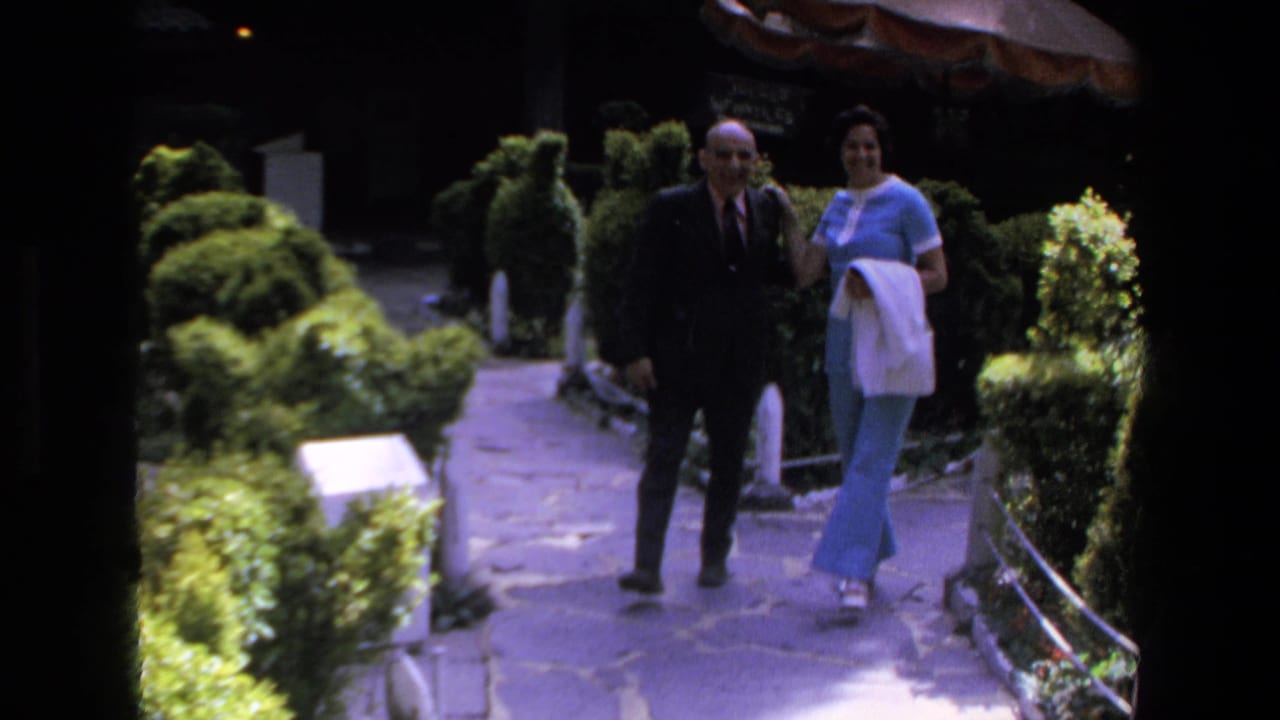 In 1972 Mazatlan home-movie footage, an older man in a dark suit and a woman in a light blue outfit walk arm-in-arm along a landscaped stone path.