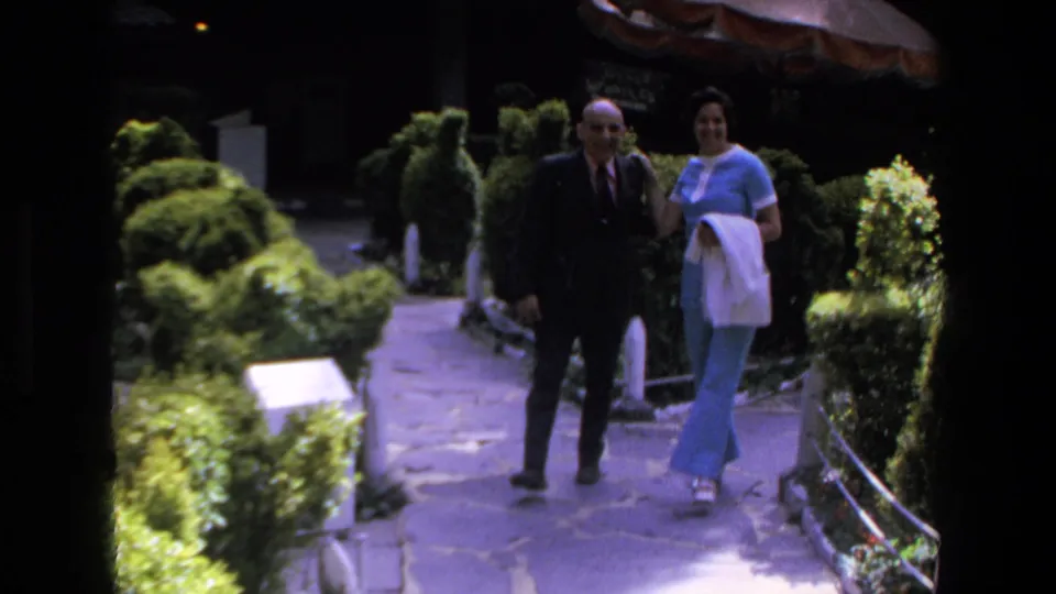 In 1972 Mazatlan home-movie footage, an older man in a dark suit and a woman in a light blue outfit walk arm-in-arm along a landscaped stone path.
