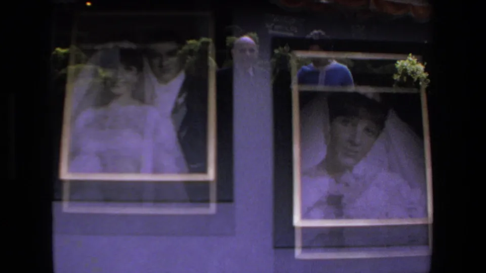 Framed wedding portraits on an indoor wall show a bride and groom, with a person faintly reflected or visible near the display in the 1972 Mazatlan clip.