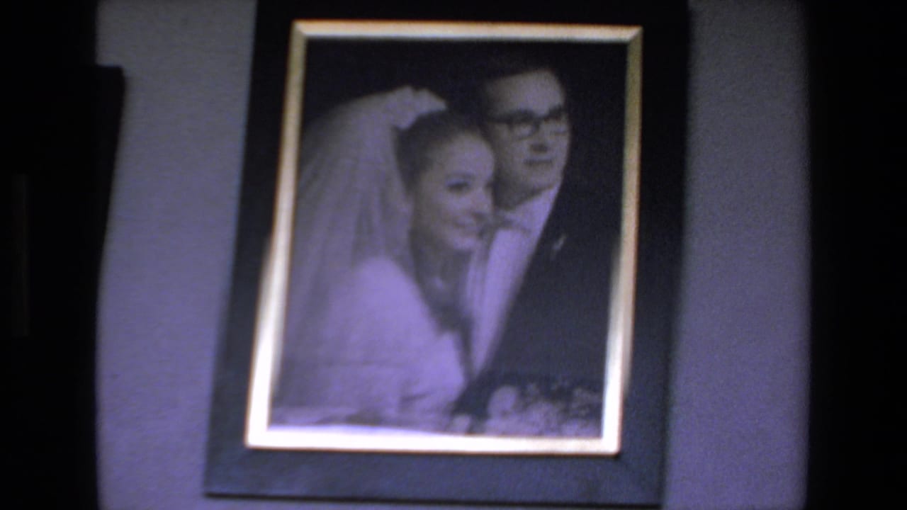 1972 Mazatlan home-movie frame showing a framed wedding portrait with a bride in a veil beside a groom in glasses, filmed in soft focus with a purple cast.