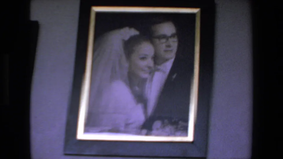 1972 Mazatlan home-movie frame showing a framed wedding portrait with a bride in a veil beside a groom in glasses, filmed in soft focus with a purple cast.