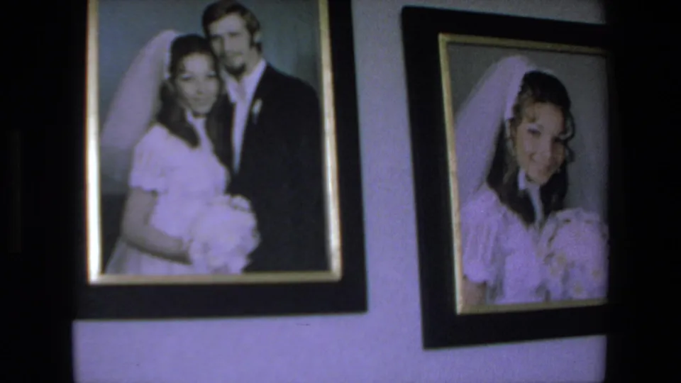 The camera frames two wedding portraits on a wall, with a bride and groom in one print and a separate bridal portrait with veil and bouquet in the other.