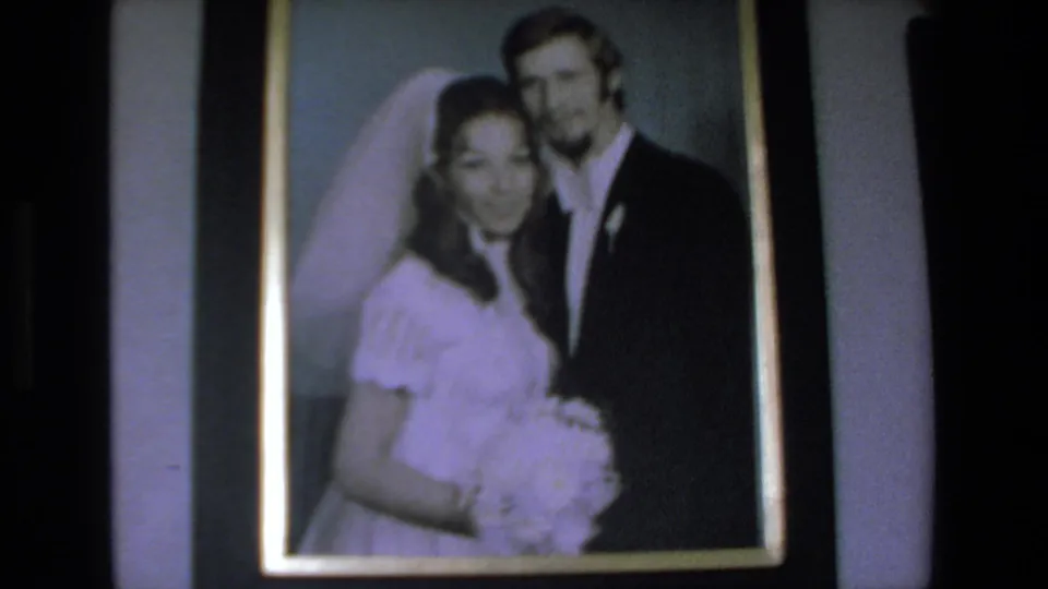 Close home-movie view of a framed wedding portrait showing a bride in a white veil and a groom in a dark suit, with soft focus and visible film grain.