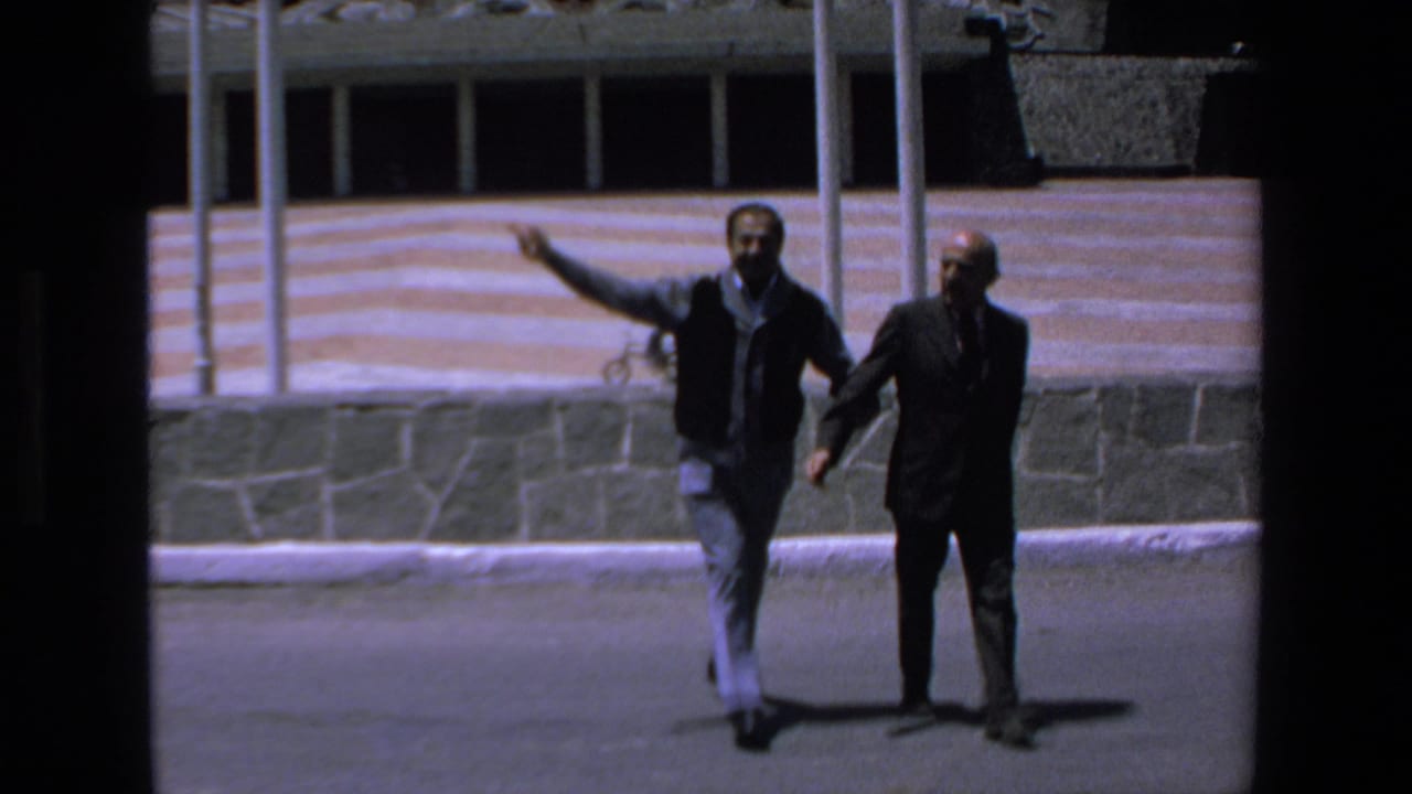 Two men walking outdoors near a low stone wall and striped facade.