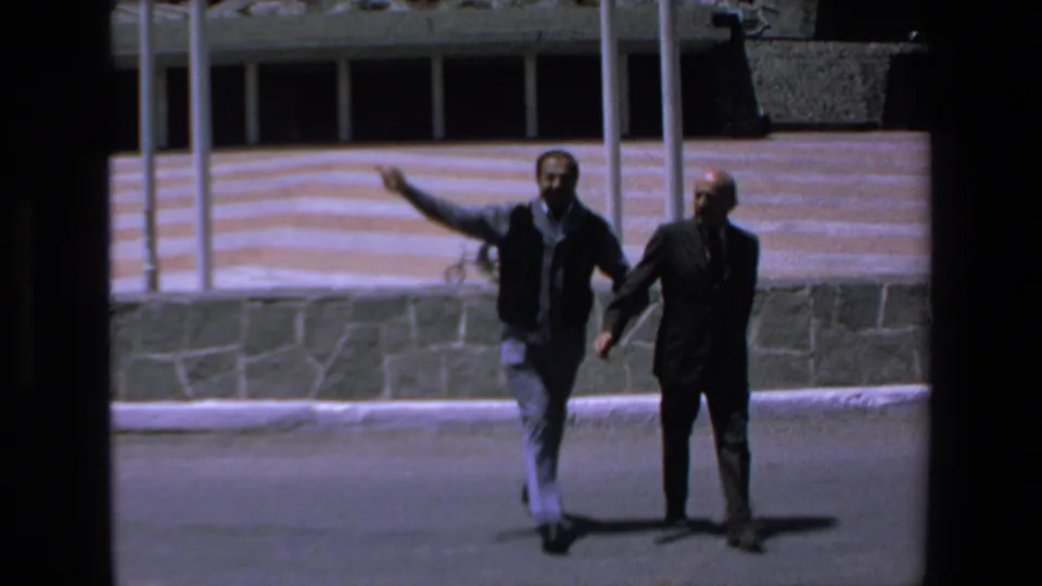 Two adult men walk across a paved outdoor area in Mazatlan, Mexico, with a low stone wall and striped building facade behind them.