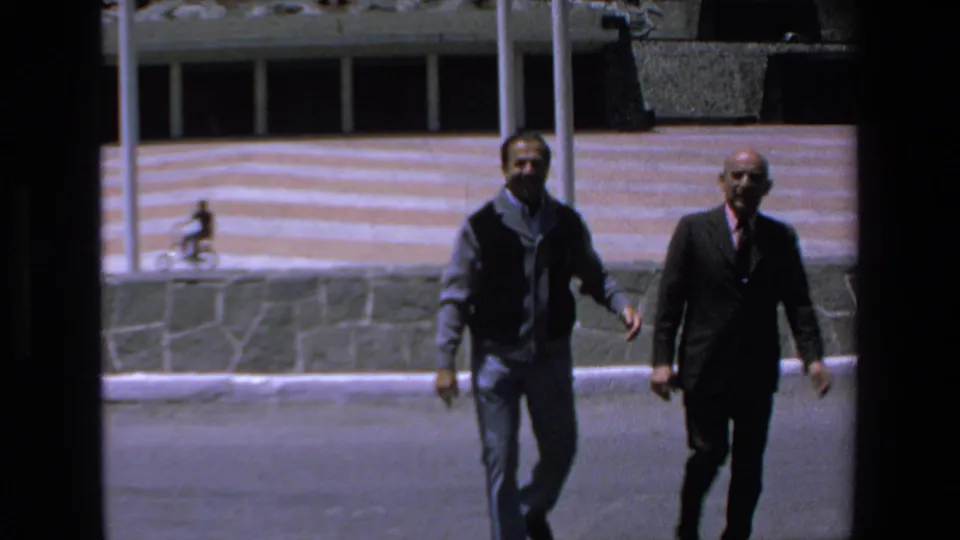Two adult men, one in casual clothing and one in a dark suit, walk toward the camera in a 1972 Mazatlan street or plaza setting.