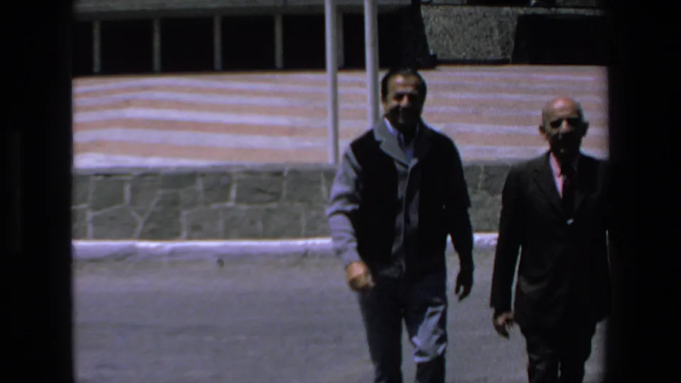 Closer 8mm frame of two adult men walking outdoors in Mazatlan, Mexico, with a low stone wall and striped facade in the background.