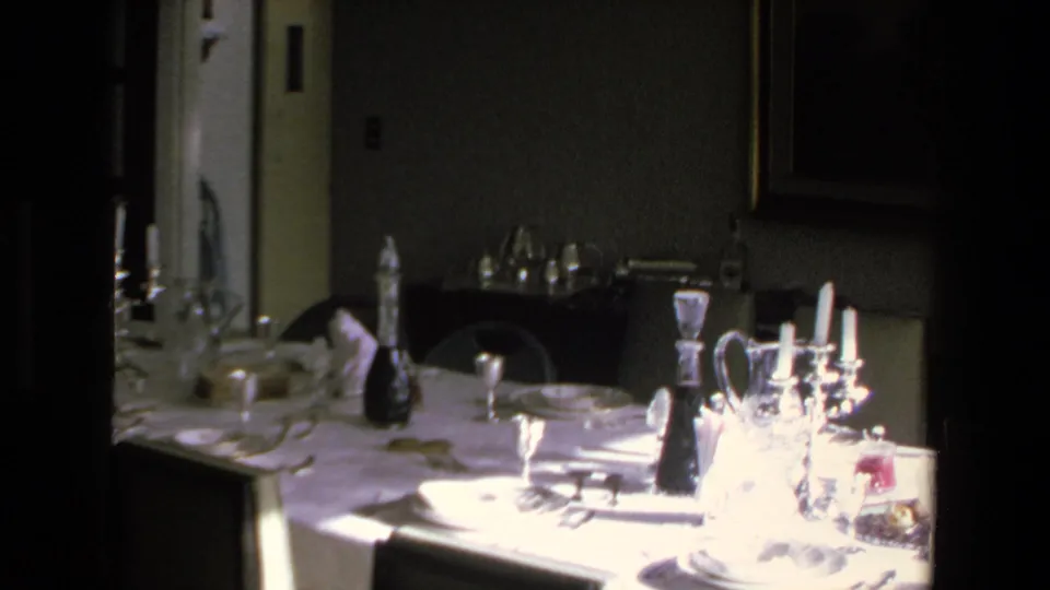 A 1972 Mazatlan home-movie frame showing a formal dinner table set with white linens, glassware, plates, and candlesticks inside a dim private dining room.