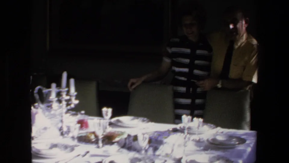 Two adults stand beside dining chairs near the formal table setting, with glassware, dishes, and bright reflected highlights visible in the 8mm frame.