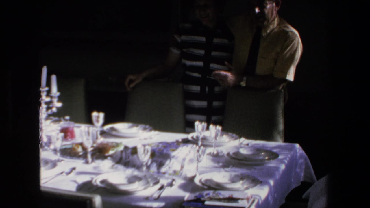 In 1972 Mazatlan home-movie footage, two adults stand beside a white-clothed dining table set with plates, glasses, and candles in a dim private interior.