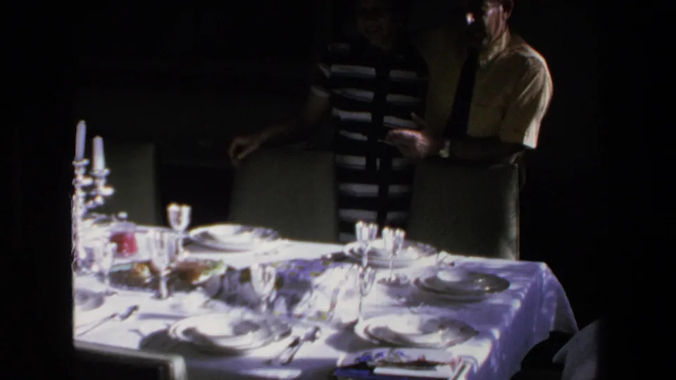 In 1972 Mazatlan home-movie footage, two adults stand beside a white-clothed dining table set with plates, glasses, and candles in a dim private interior.
