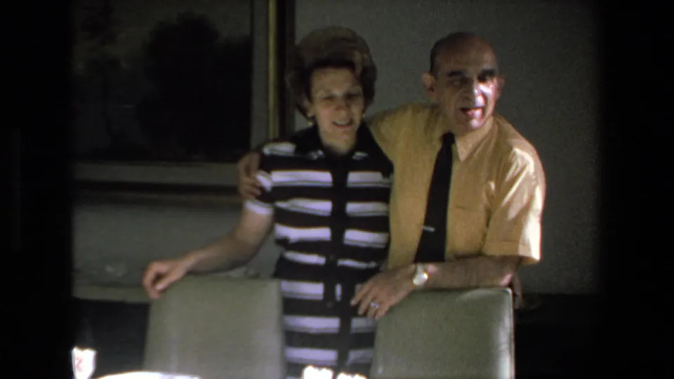 The pair stand close together behind dining chairs, with the formal table setting and framed wall art partly visible in the 1972 Mazatlan home-movie frame.