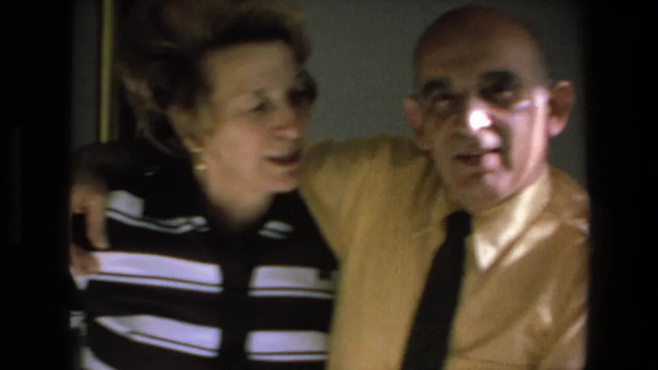 In 1972 Mazatlan home-movie footage, an elderly couple sit closely together indoors as the woman in a black-and-white striped top leans toward the man in a yellow shirt and dark tie.